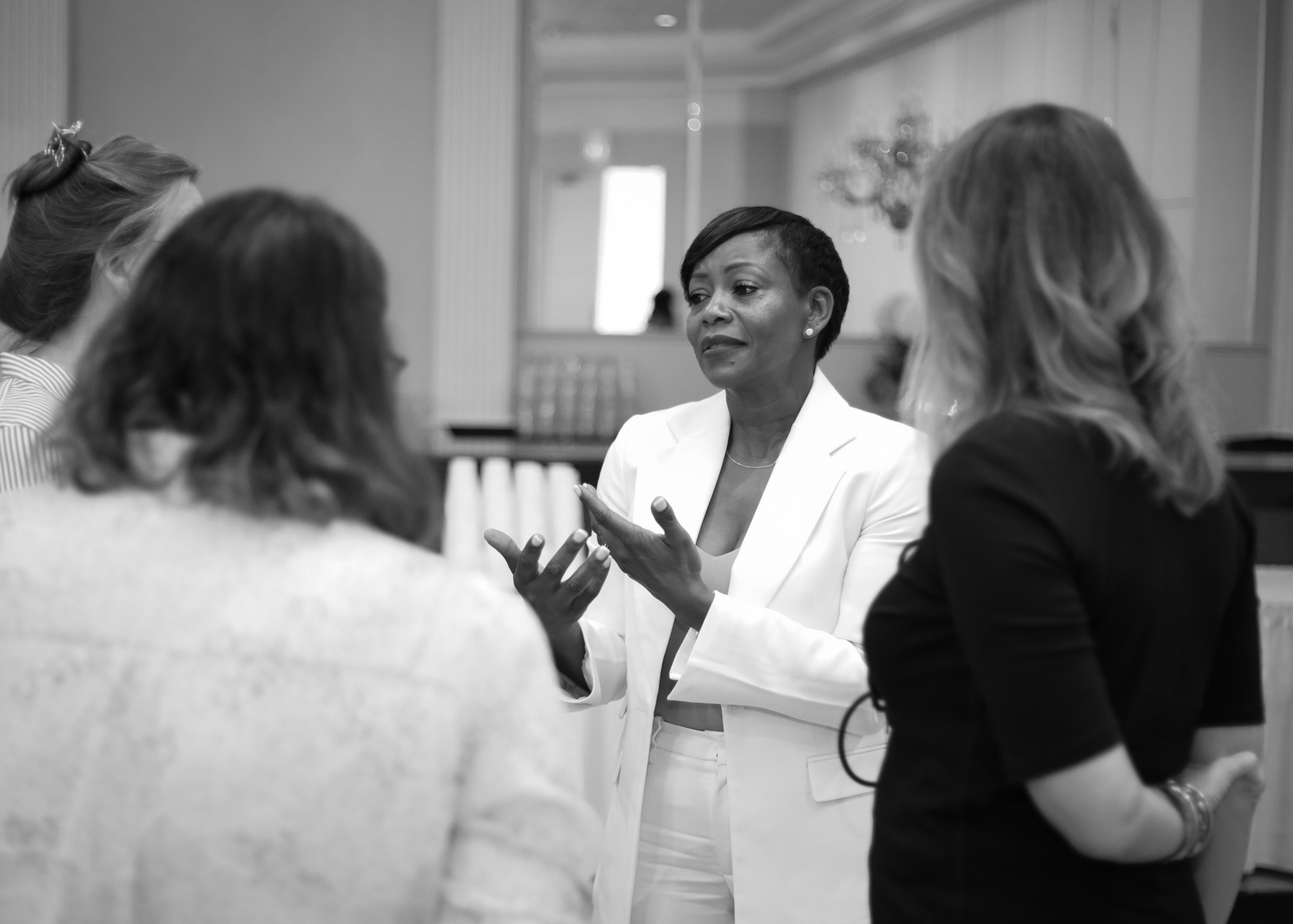 Woman in white suit gesturing, speaking to group indoors.