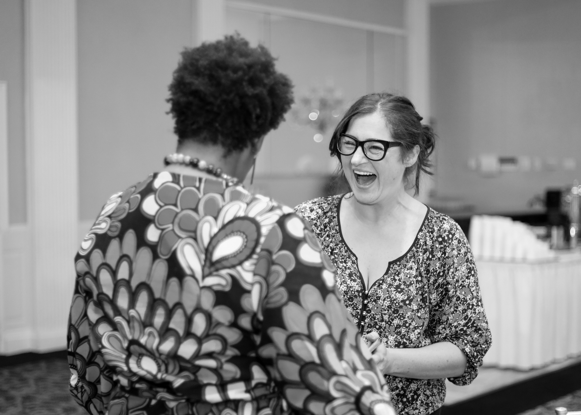 Woman laughs while talking to another person wearing a patterned shirt. Indoor setting with a buffet table.