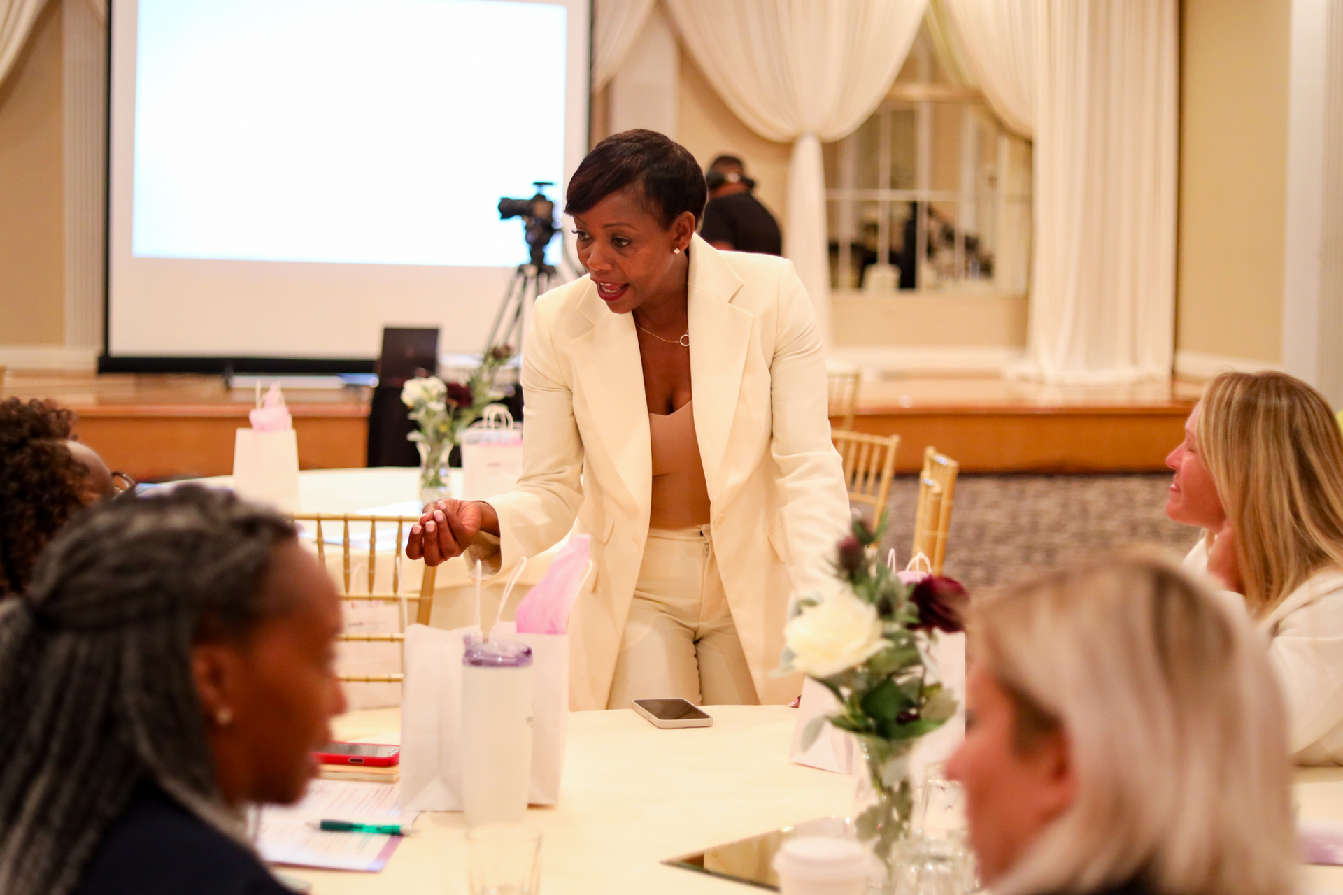 Woman in cream suit speaks at a table to other women in a conference room.