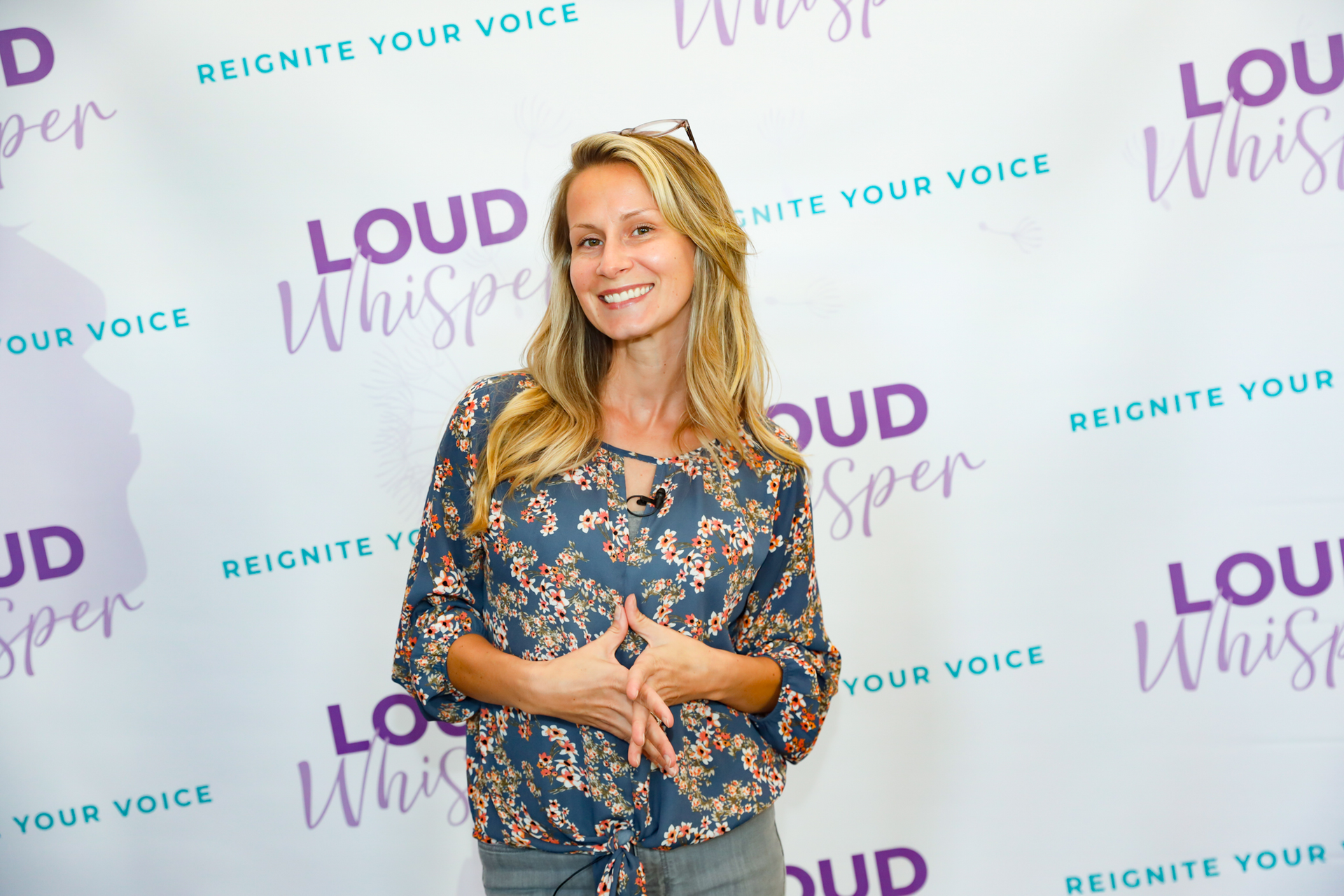 Woman in floral top smiles, hands clasped, in front of a backdrop reading 