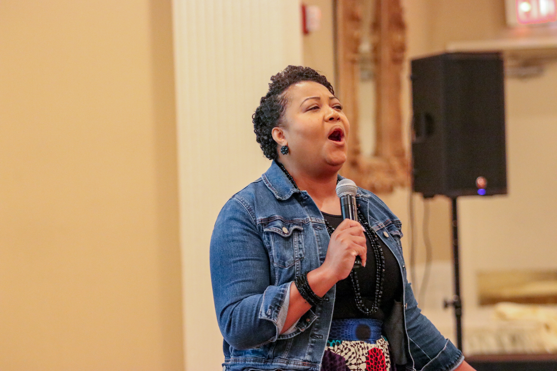 Woman singing into a microphone, wearing a denim jacket, in a venue.
