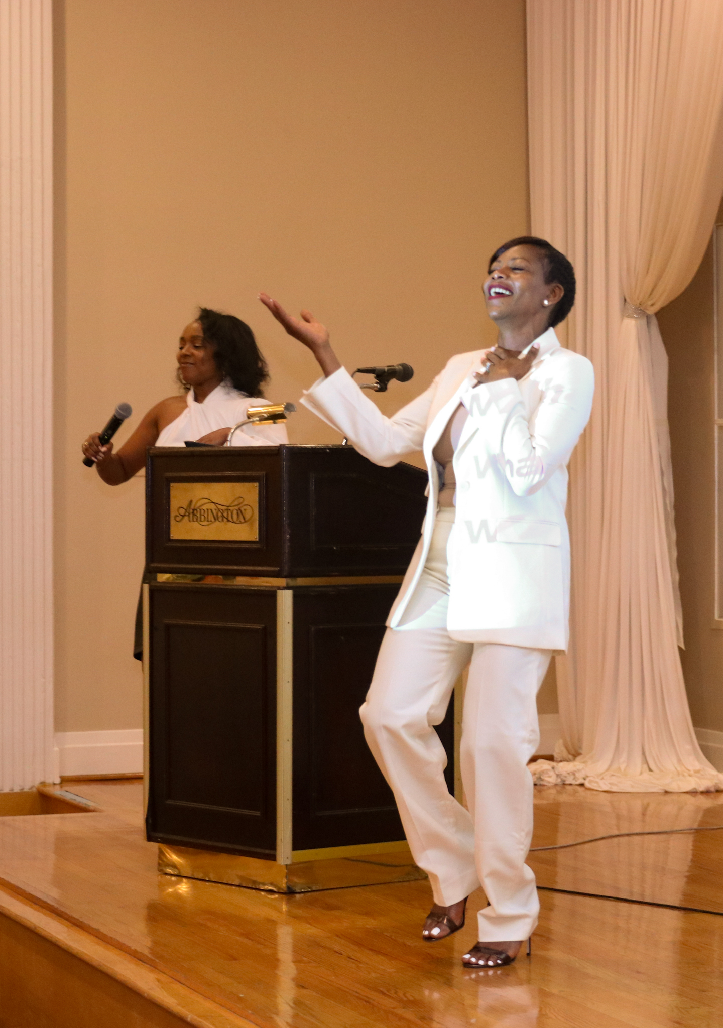 Woman in white suit singing on stage, another woman behind a stand with a microphone. Wooden floor.