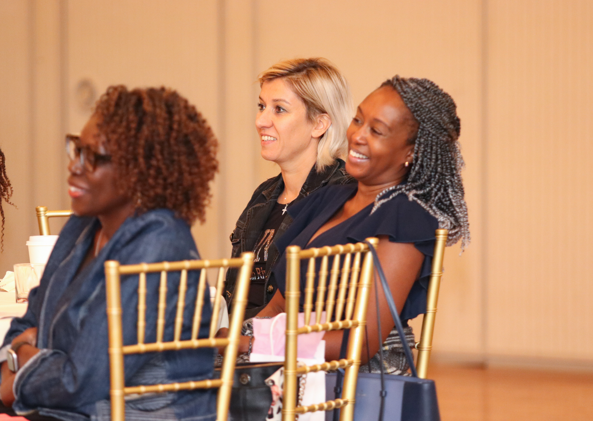 Three women smiling, seated in golden chairs, in a bright room.