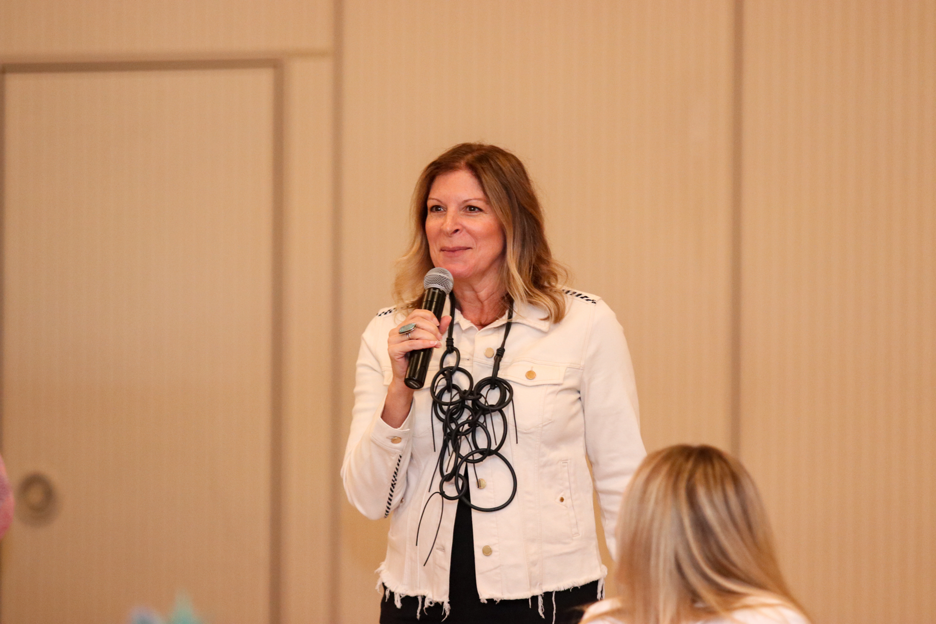 Woman speaking into microphone, wearing white jacket, standing in front of light beige wall, and another woman.