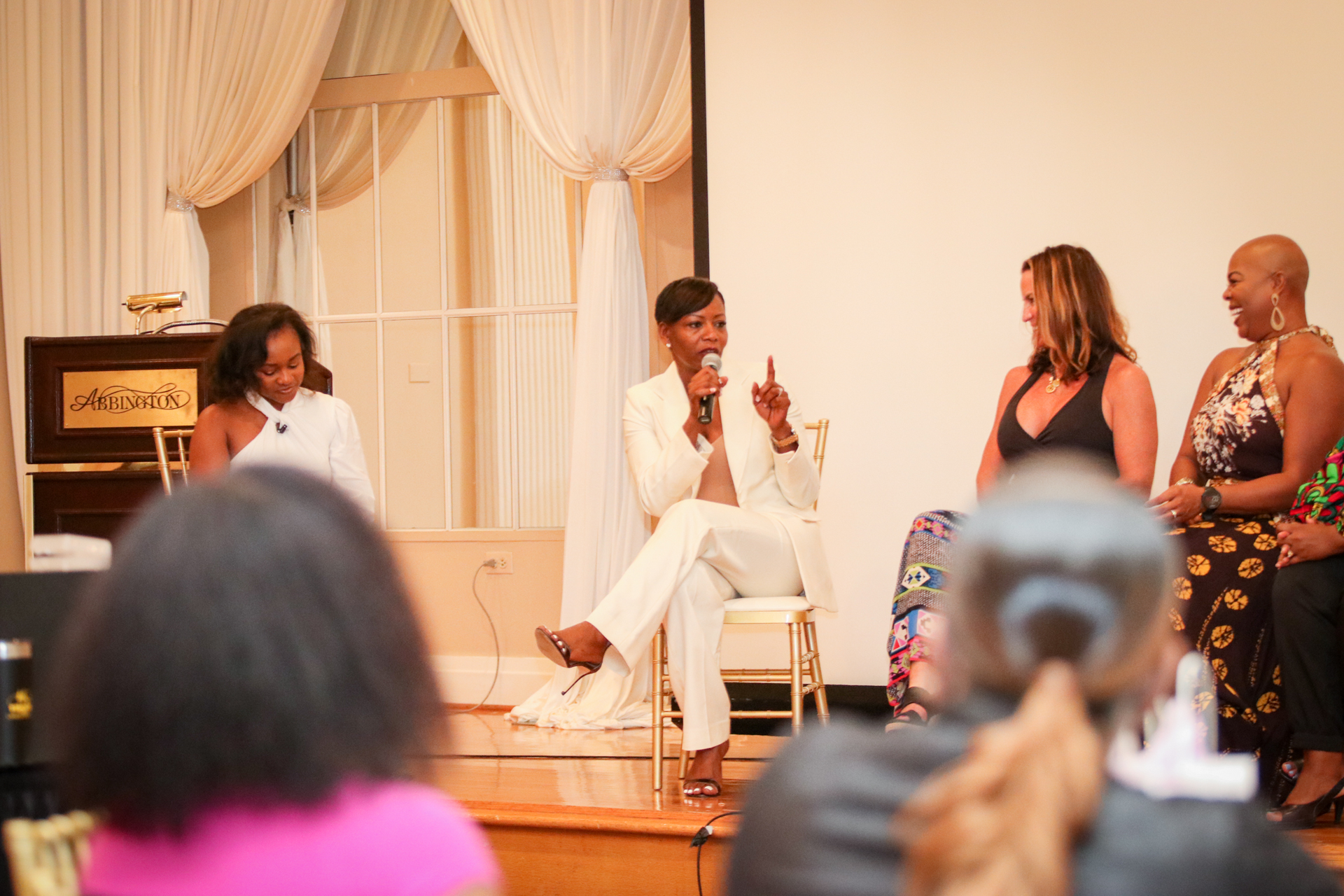 Woman in white suit speaks on stage during a panel. Other women stand or sit, audience in foreground.