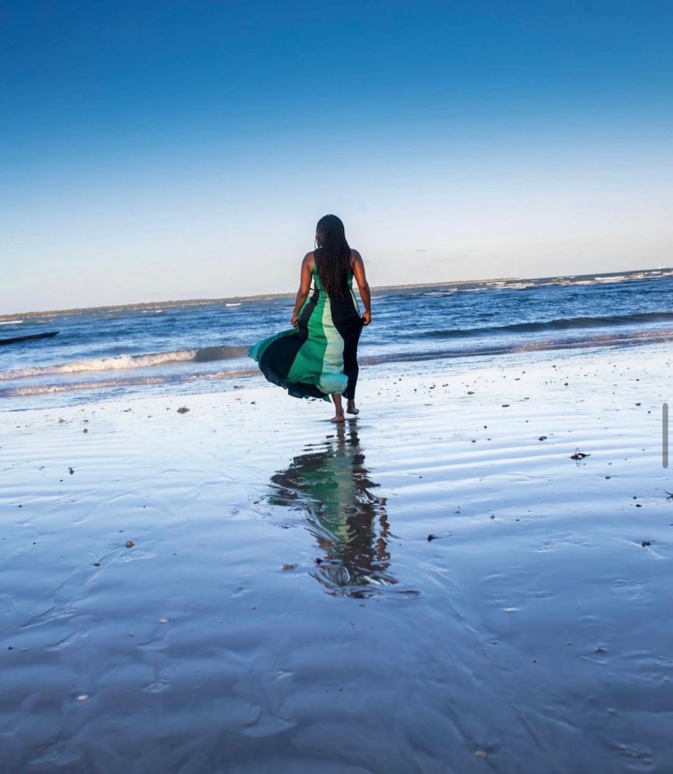 Person in a flowing green dress walking on a wet beach with ocean waves and blue sky reflected in the sand.