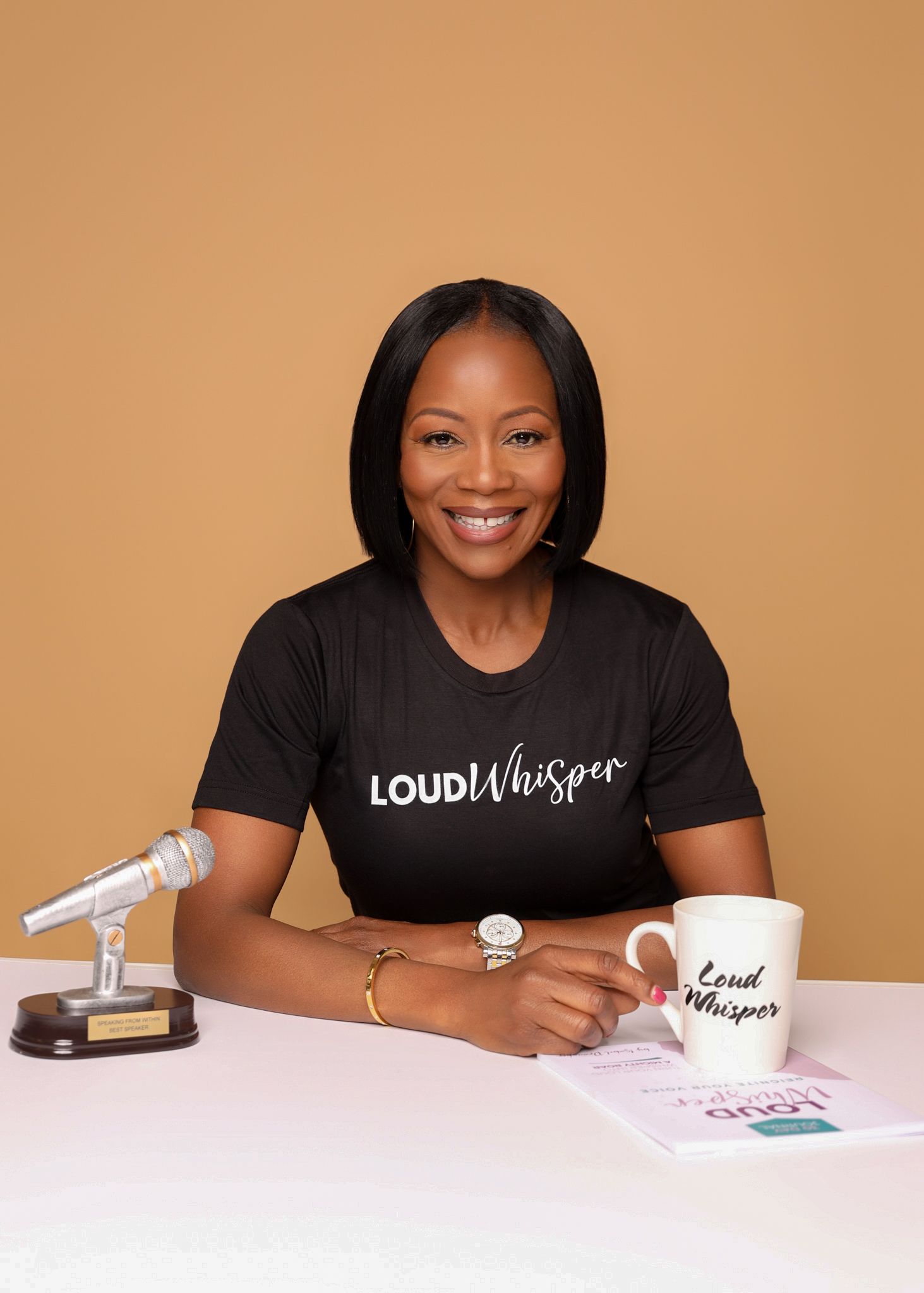 Woman smiling at a desk with a mug, microphone, and black T-shirt against a tan background.