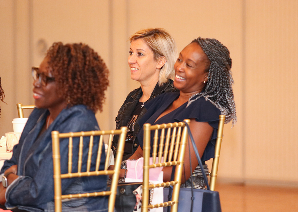 Three women smiling while seated; indoor event, golden chairs, beige walls.