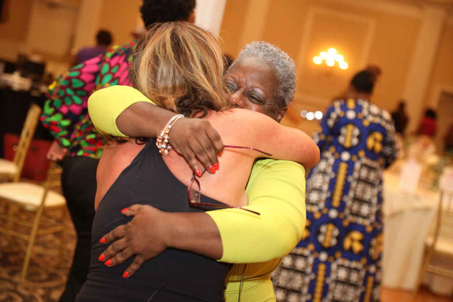 Two people embracing indoors; one in black top, the other in yellow. Warm smiles, room setting.