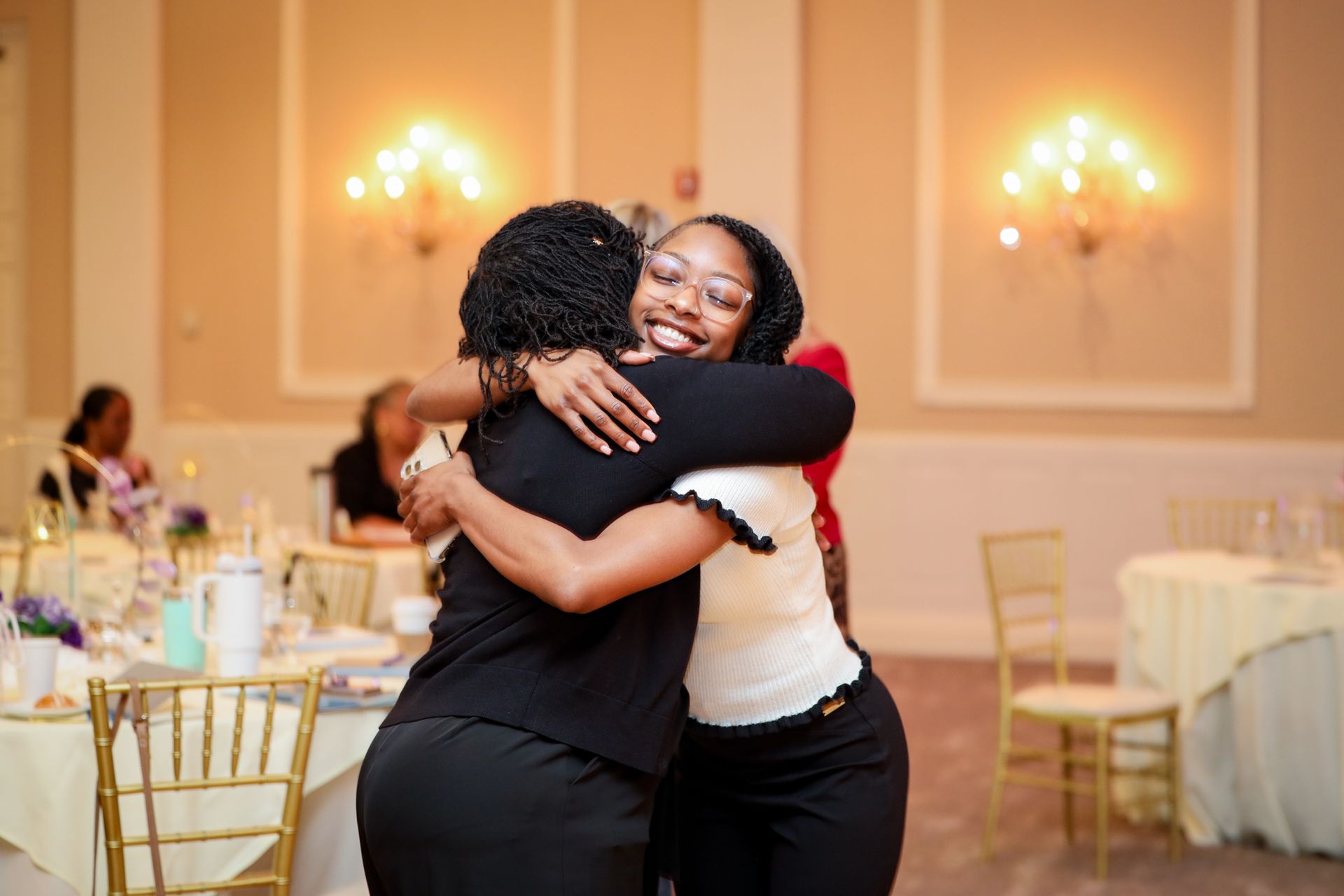 Two people hugging in a warmly lit banquet hall with round tables and chairs