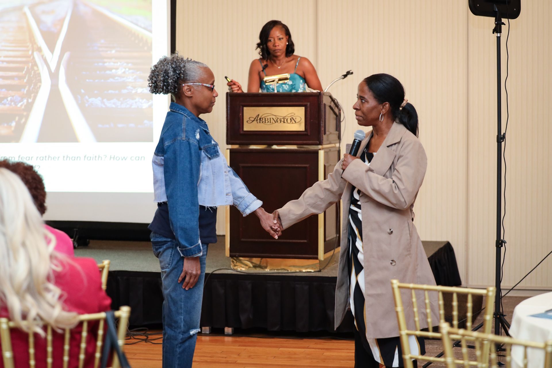 Two people shake hands at a podium during an indoor event, with a speaker in the background.