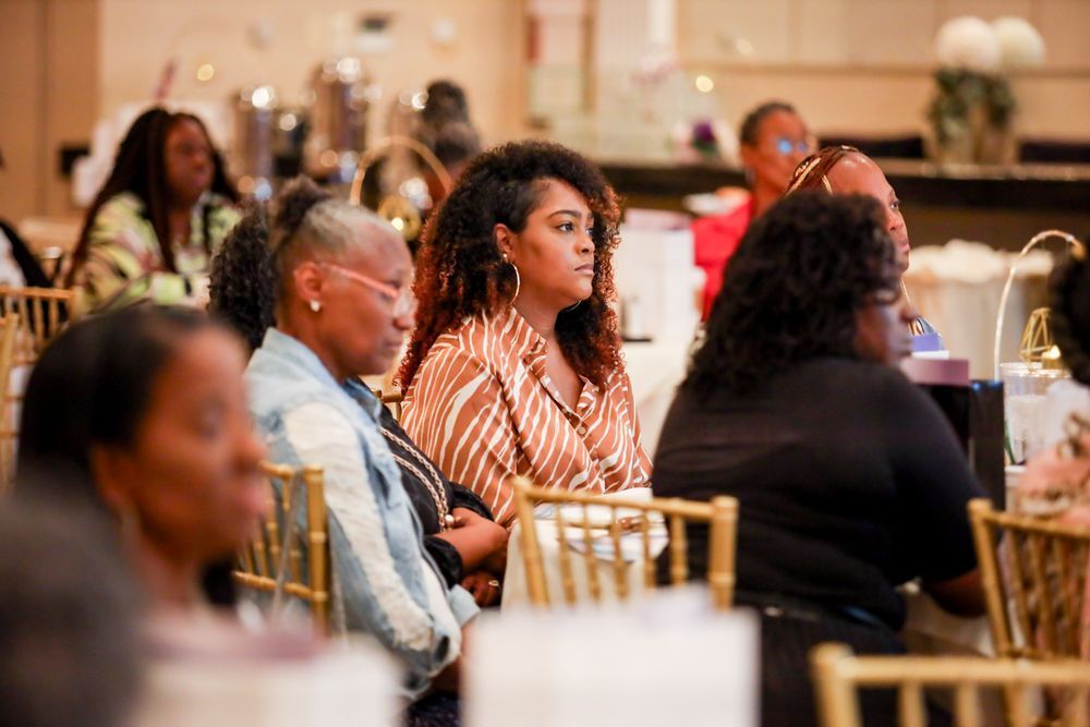 Attendees at an event seated at tables, listening. The room has soft lighting.