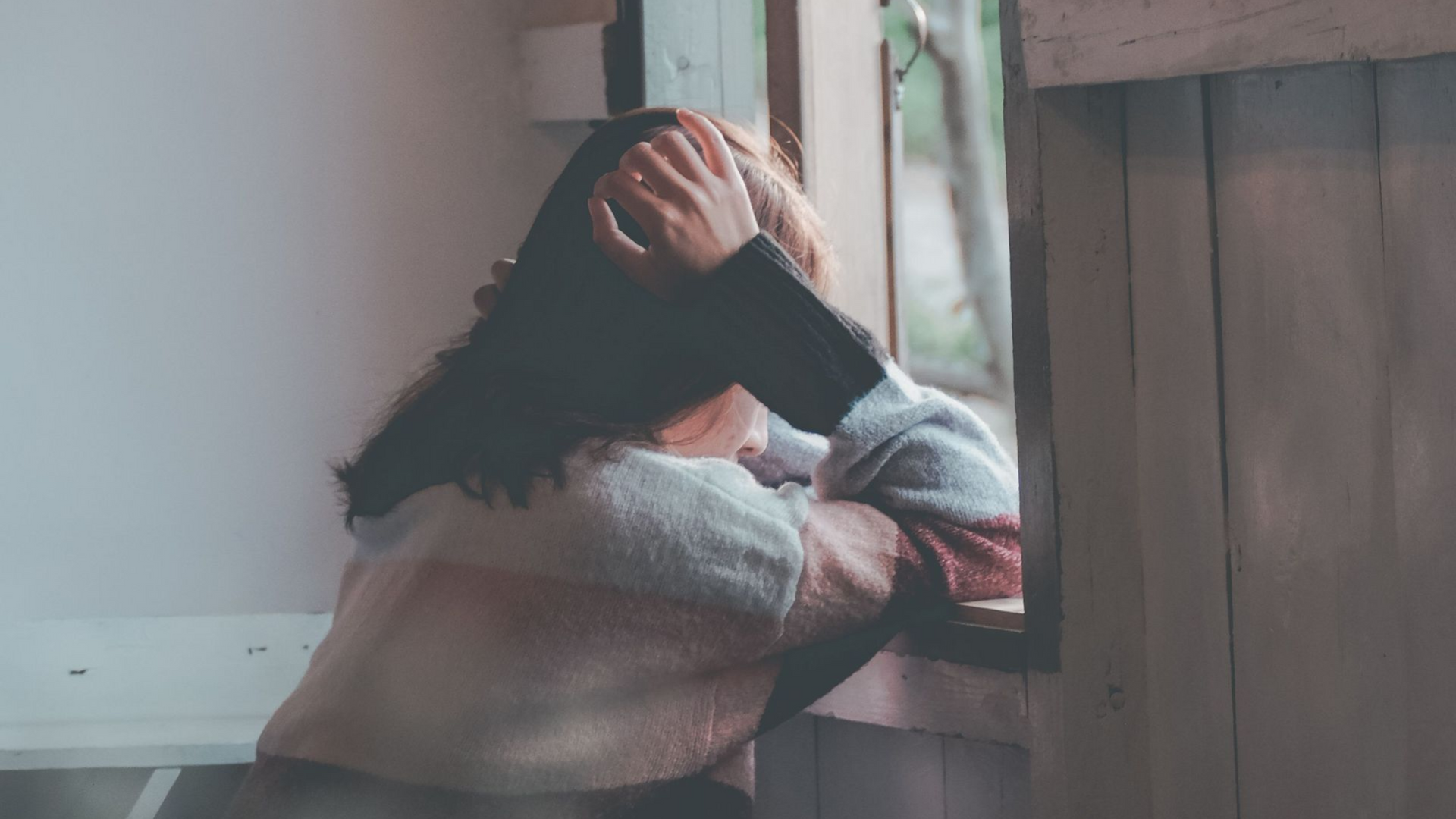 Person sitting by a window with head in hands, looking distressed in soft natural light