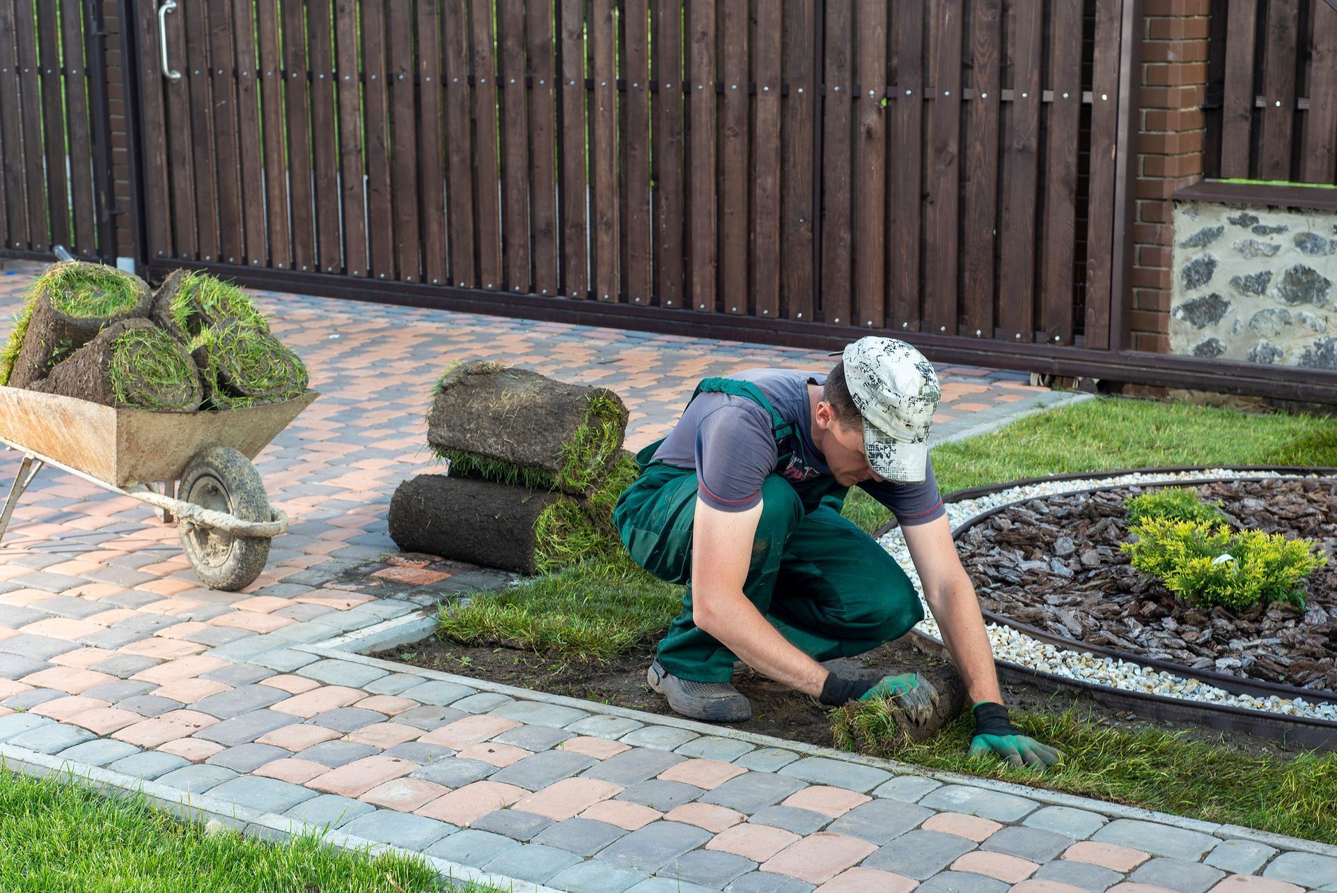 A man is kneeling down in a garden with a wheelbarrow full of grass.
