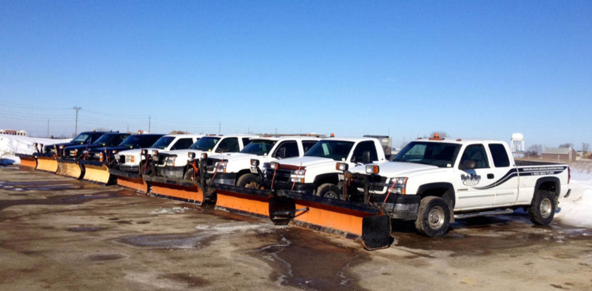A row of snow plows are parked in a parking lot.