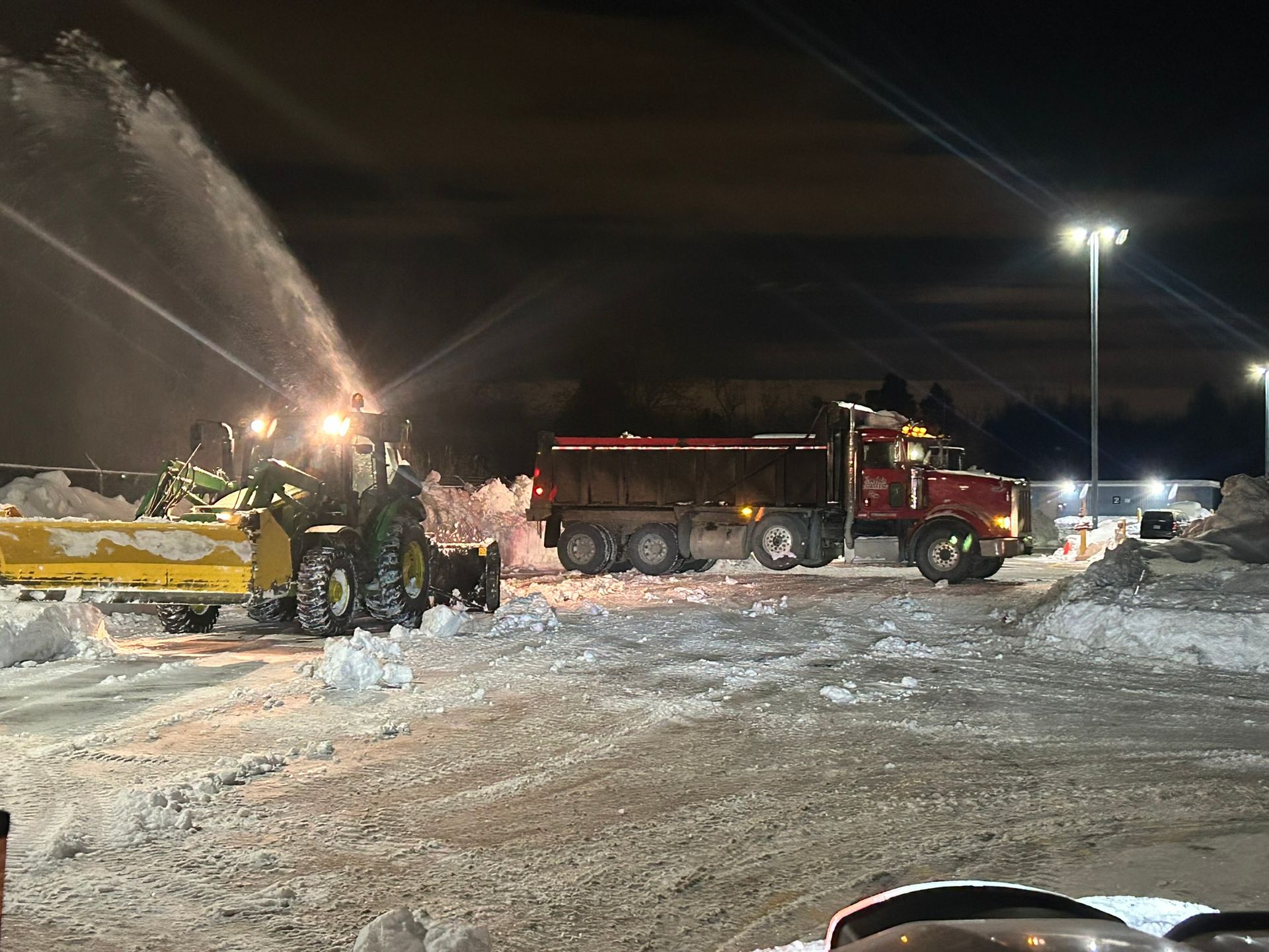 A snow plow is blowing snow next to a dump truck at night.