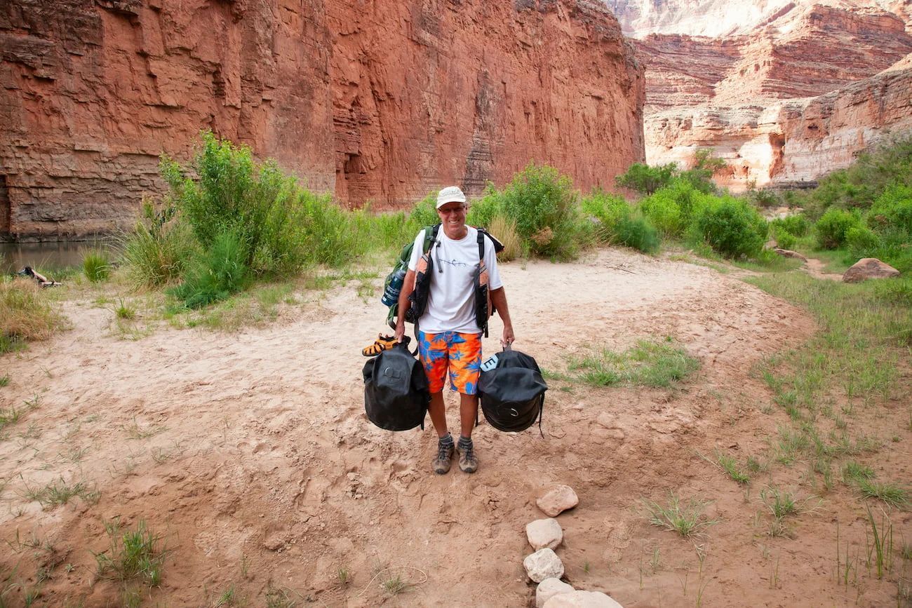 Man hikes along a sandy path, carrying two black bags and a backpack, canyon in background.