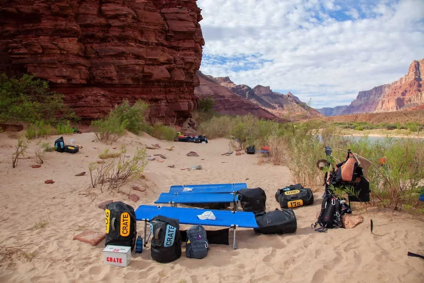 Camping gear on a sandy beach near a red rock cliff and a river under a cloudy sky.