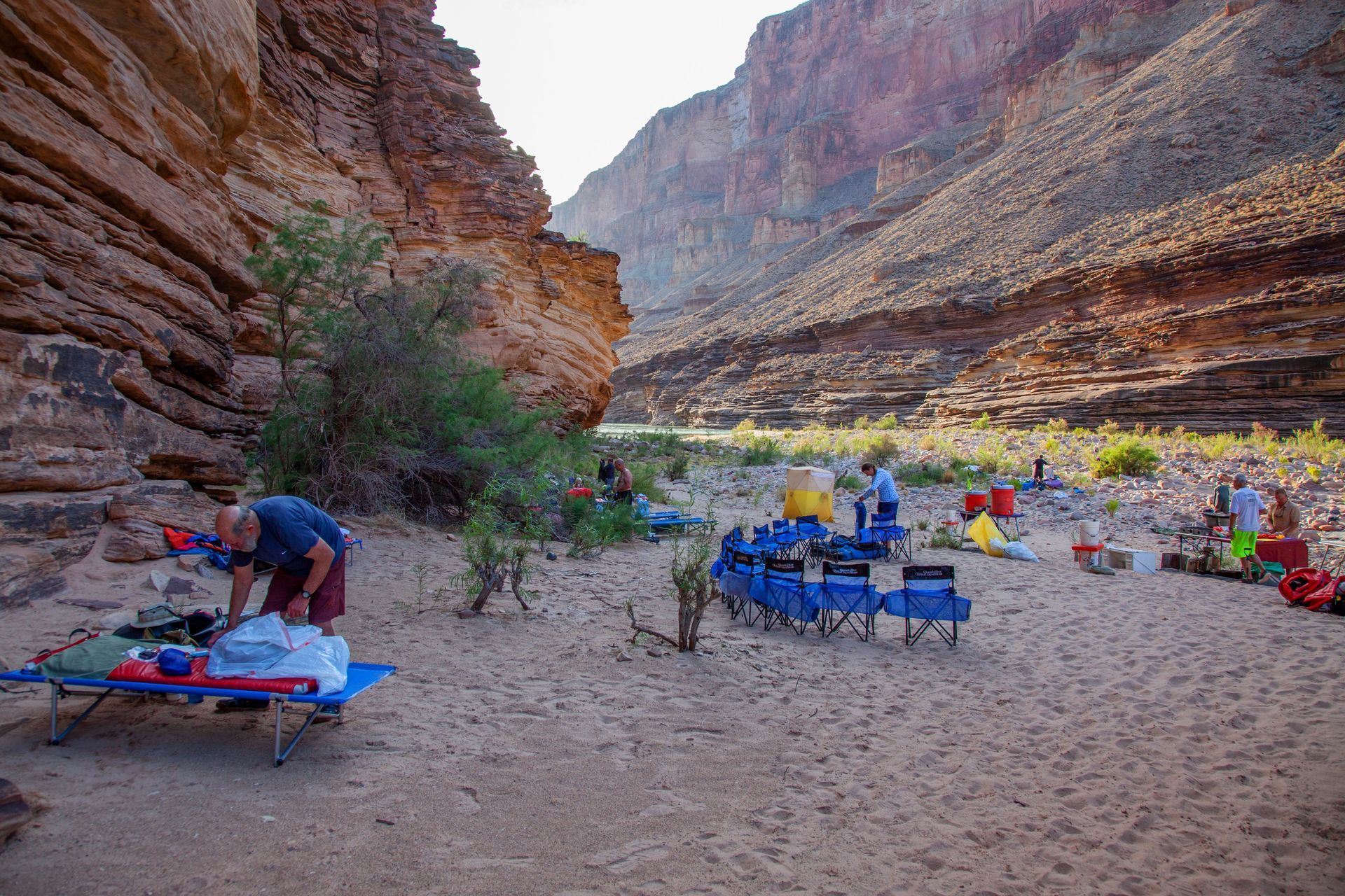 Campers set up on a sandy beach at the base of a canyon. Blue chairs and gear are scattered.