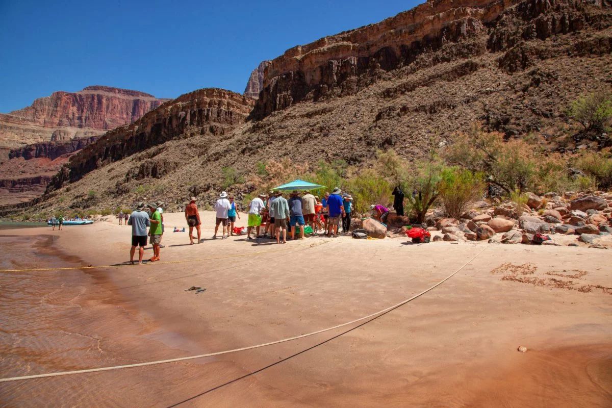 People gathered on a sandy beach next to a river with canyon walls in the background under a sunny blue sky.