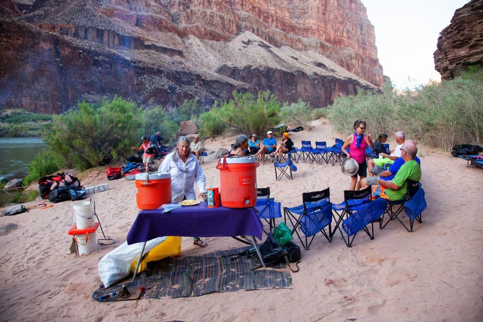 People seated on folding chairs gather on a sandy riverbank, preparing food with a canyon backdrop.