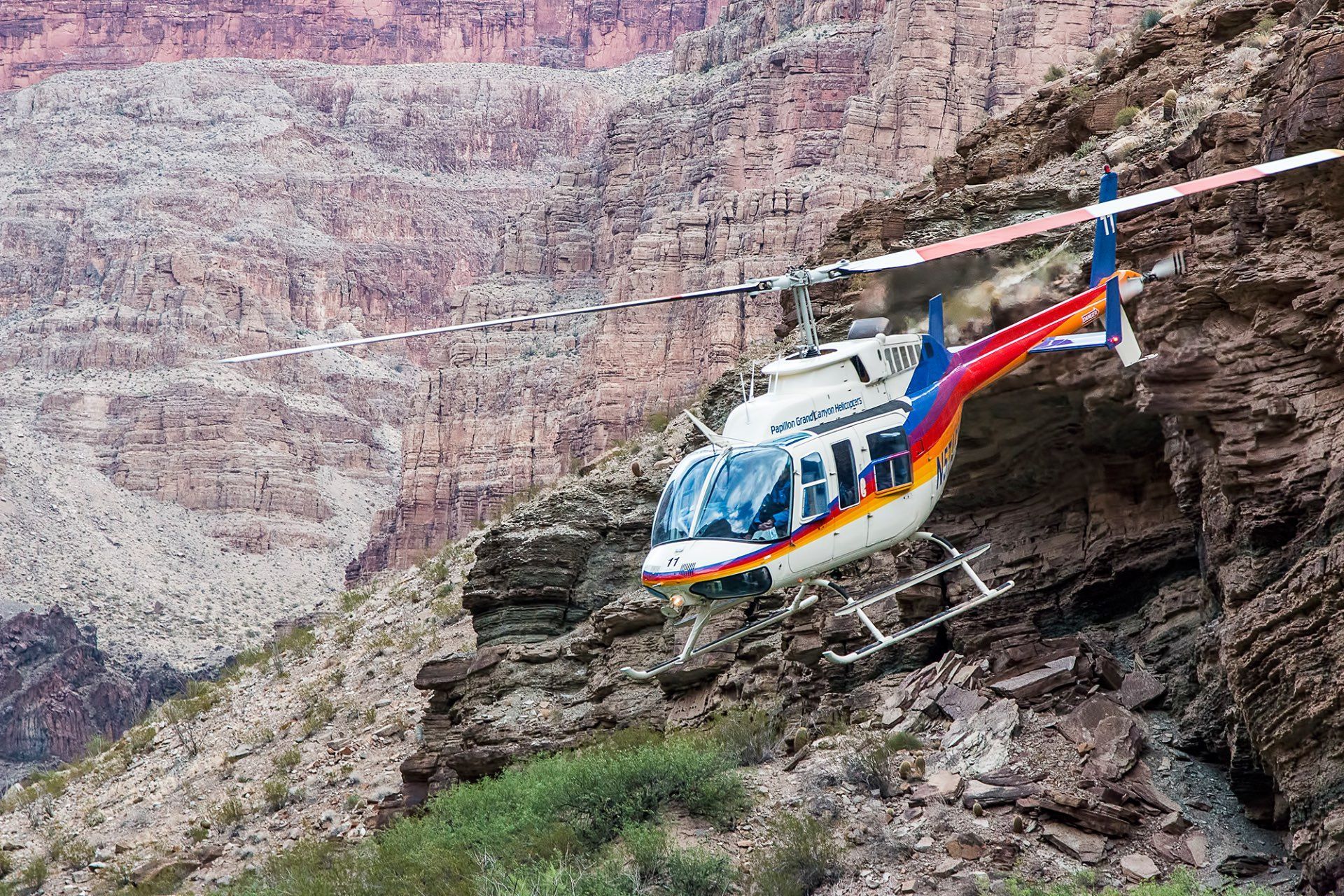 Helicopter flies near a rock face in the Grand Canyon. It is white, red, yellow, and blue.