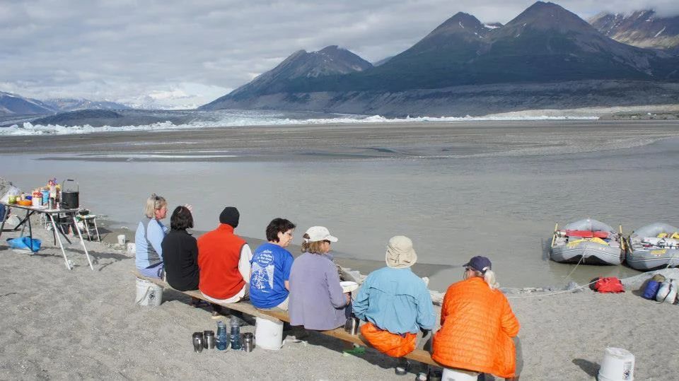 People seated on a bench by a river, mountains in the background. Rafts and gear are nearby.