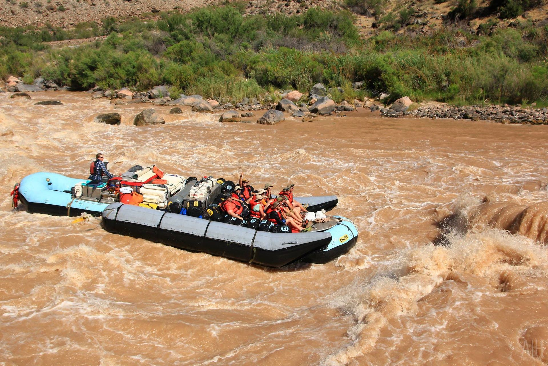 Raft with passengers navigates a rapid in a muddy river, surrounded by rocky landscape and greenery.