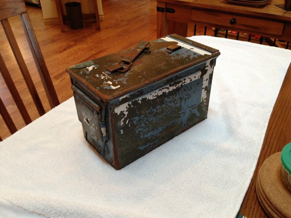 A weathered, olive-drab metal box with a latch sits on a white towel on a wooden table.