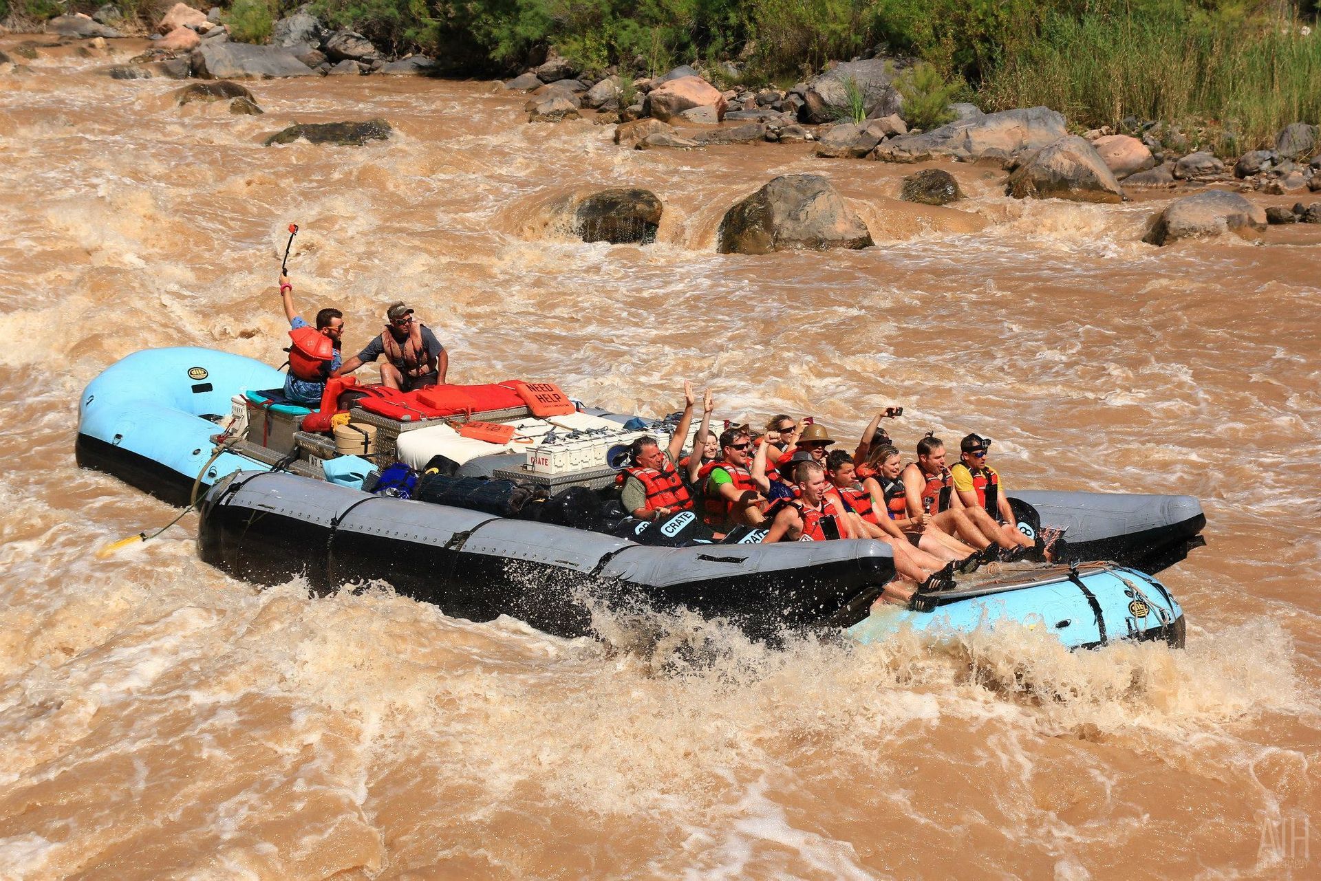 Raft of people on a rushing brown river. They wear life vests, smiling and waving.