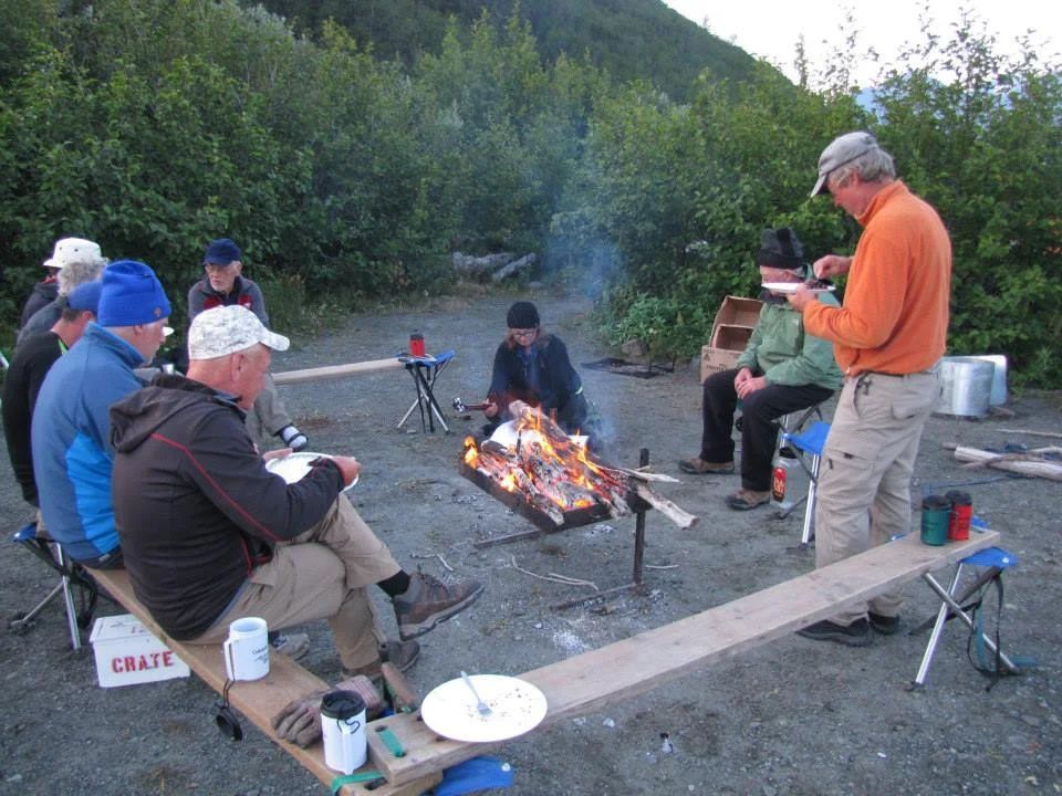 People around a campfire, eating. Men seated on benches, near trees, dusk setting.