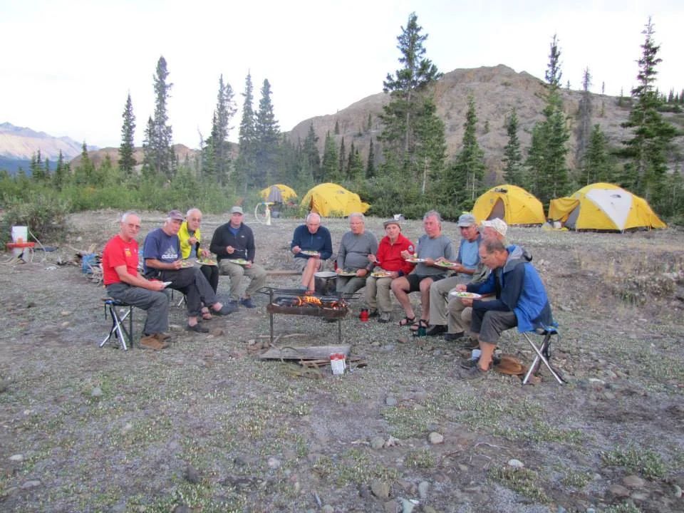 A group of men camping, gathered around a small fire, with tents and mountain scenery in the background.