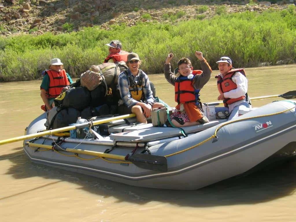 People on a raft on a river, smiling and cheering. Green foliage in the background.