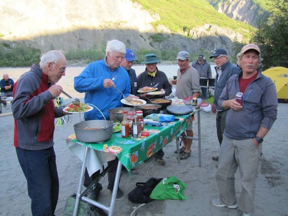 Group of people at an outdoor meal, serving themselves from various dishes on a table; river and mountains in the background.