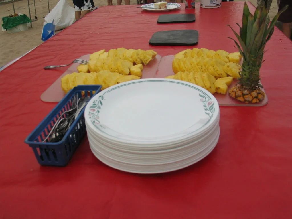 Table with sliced pineapples, plates, and utensils on a red tablecloth.