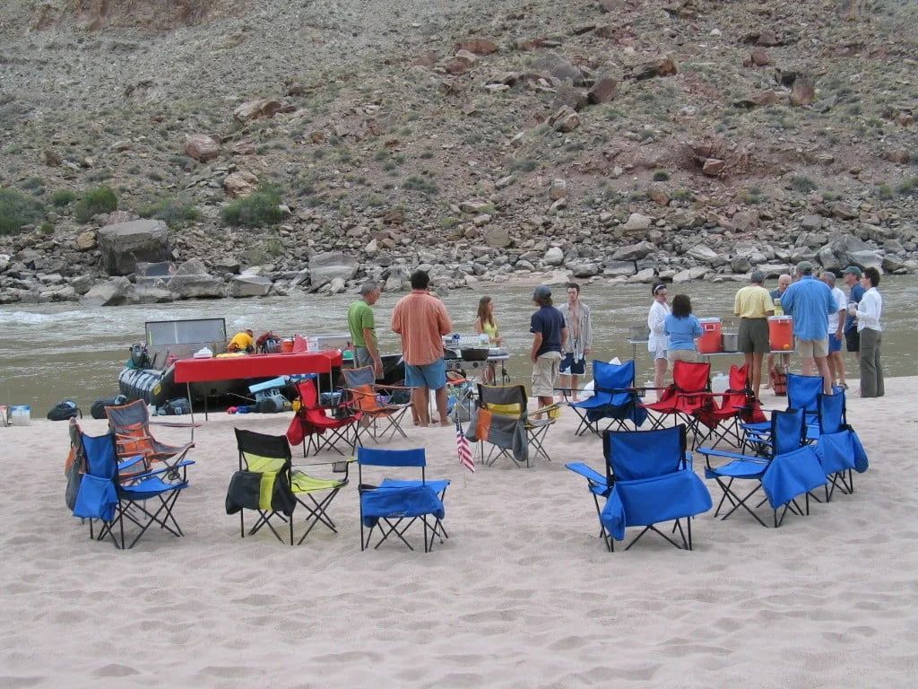 Group of people by a river gathered around chairs and a table on a sandy shore.