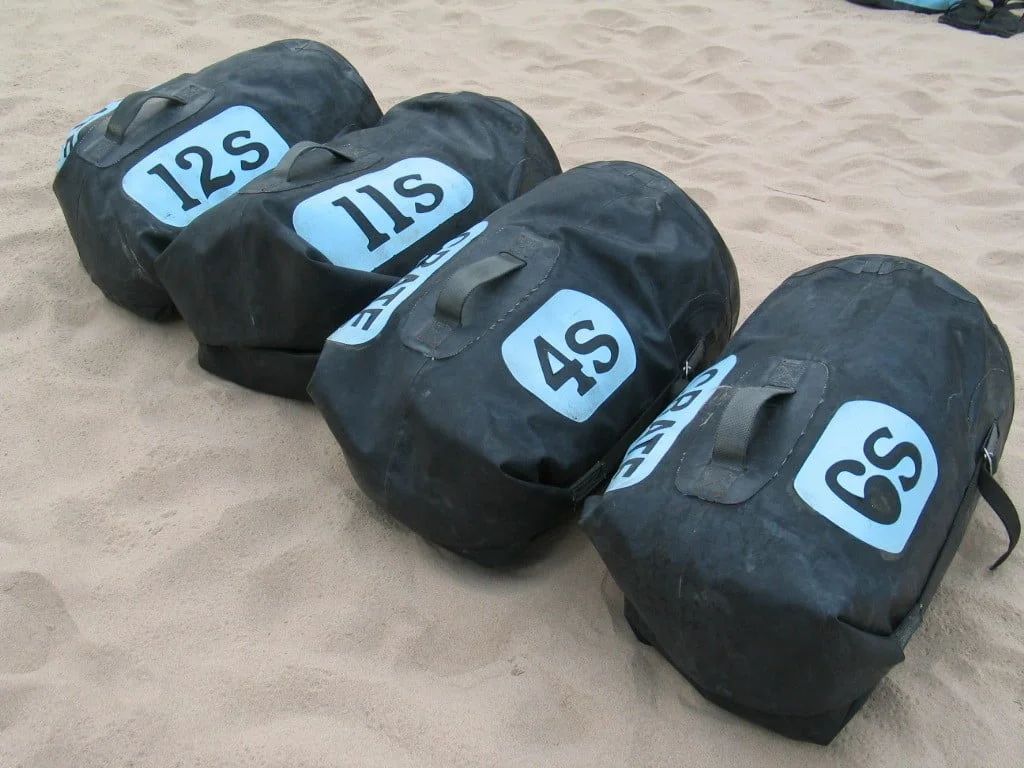 Four black sandbags with blue labels marked 12S, 11S, 4S, and 8S, placed on sand.