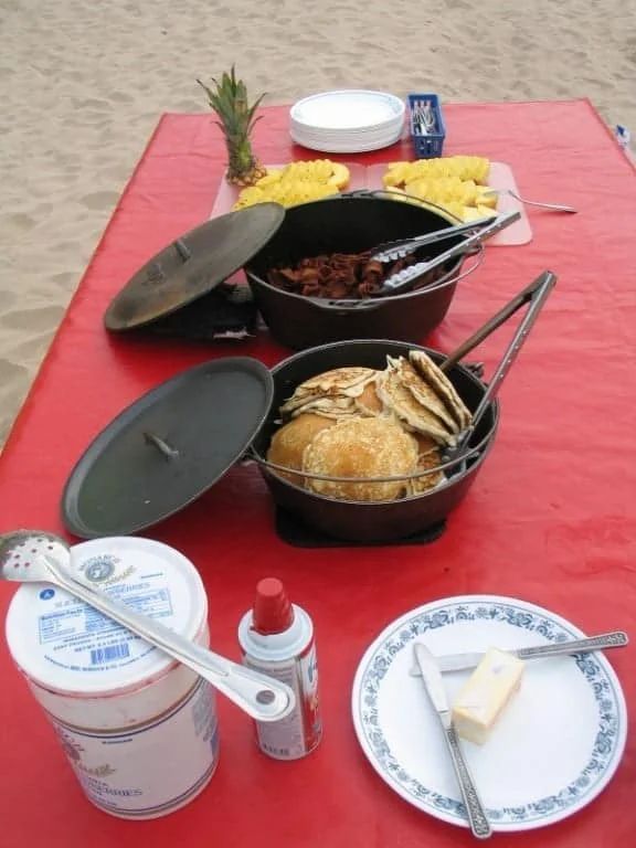 Food on a red table: Dutch ovens, corn, pineapple, butter, whipped cream, and utensils.