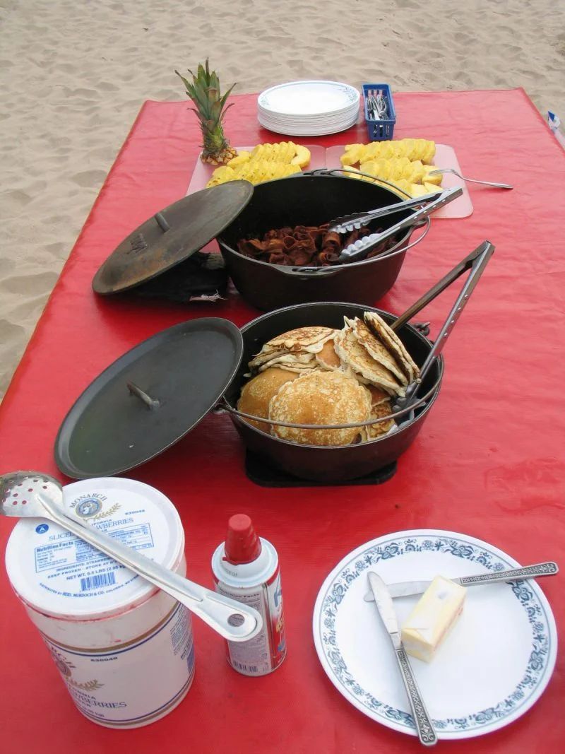 Picnic on the beach: cast iron pots with food, pineapple, corn, bread, butter, and condiments on a red tablecloth.