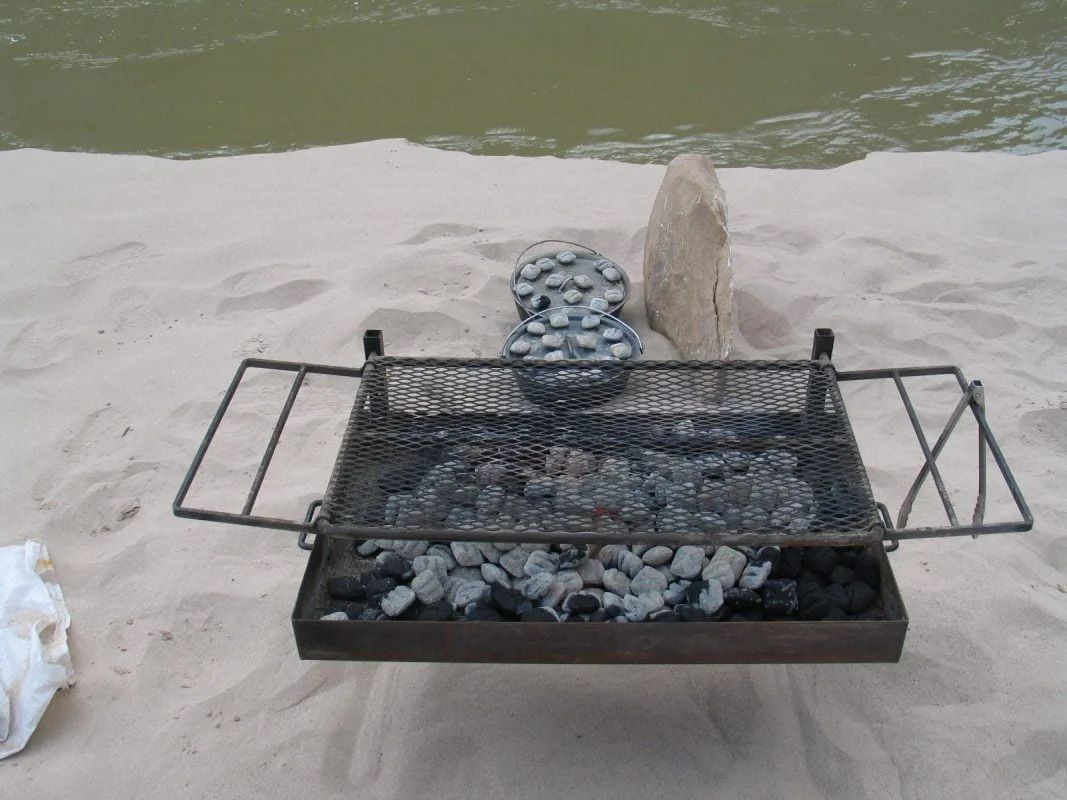 Charcoal grill on sandy beach with water in the background, near a large rock.
