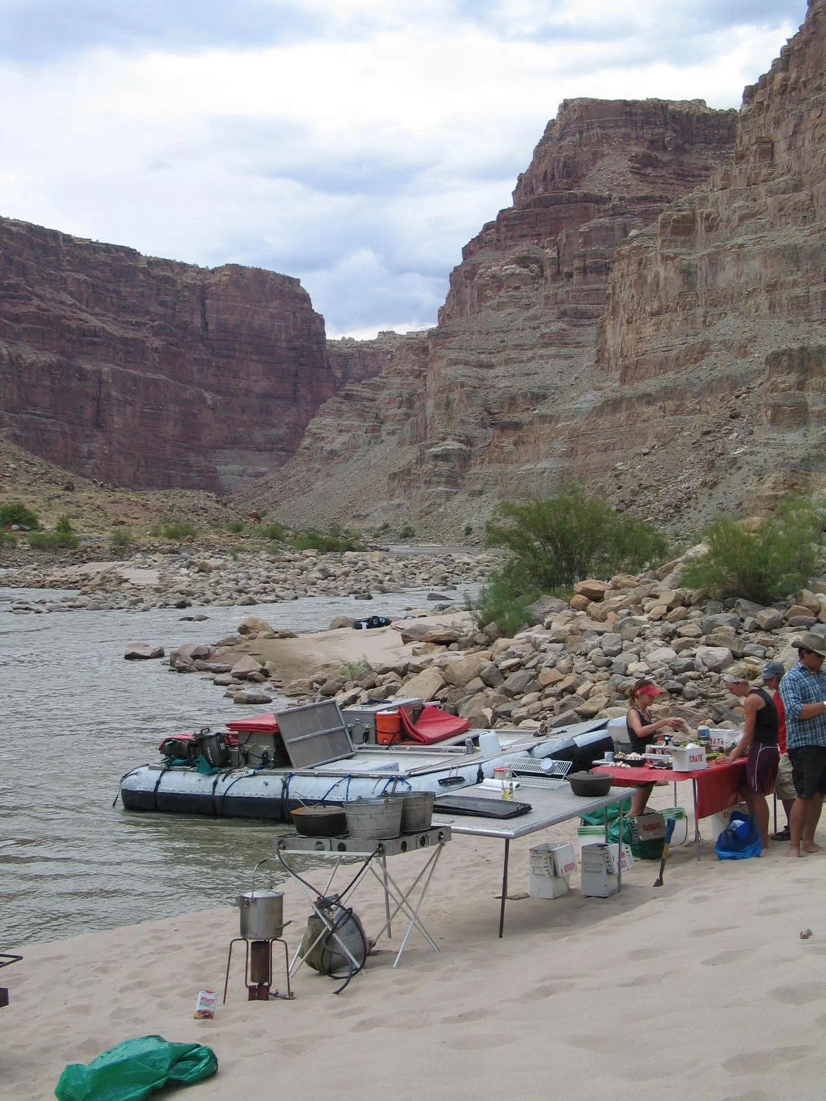 Riverbank campsite with raft, cooking setup, and people; red rock canyon backdrop.