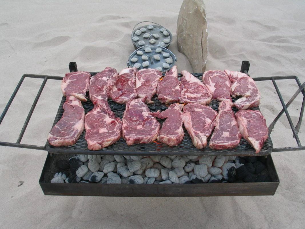 Lamb chops grilling on a metal grate over charcoal on a sandy beach.