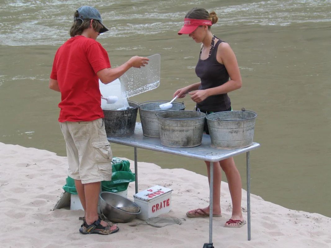Two people serving something from buckets on a table near a river.