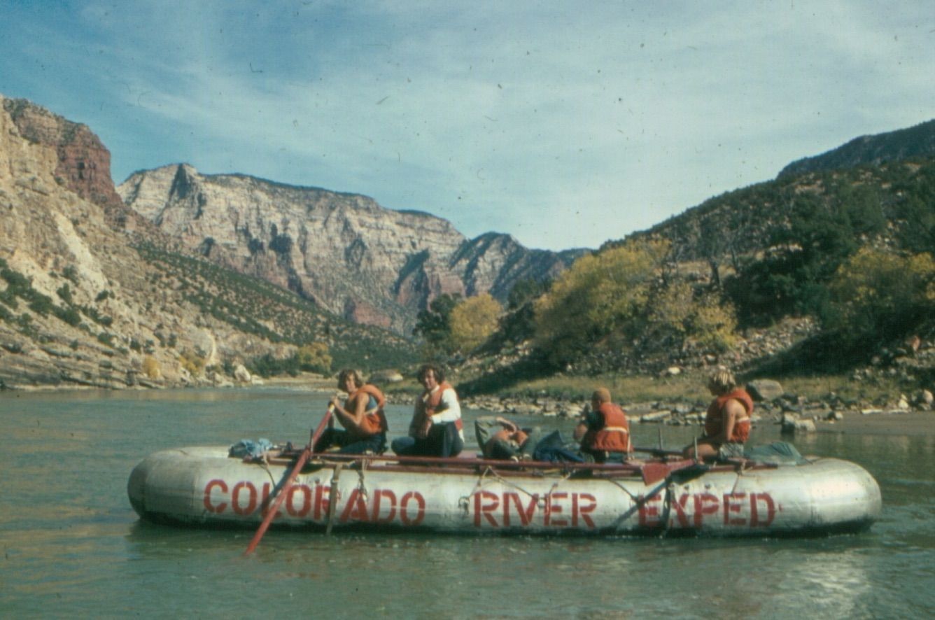 Rafting on Colorado River. People in orange life vests row an inflatable boat past canyon walls. Sunny day.