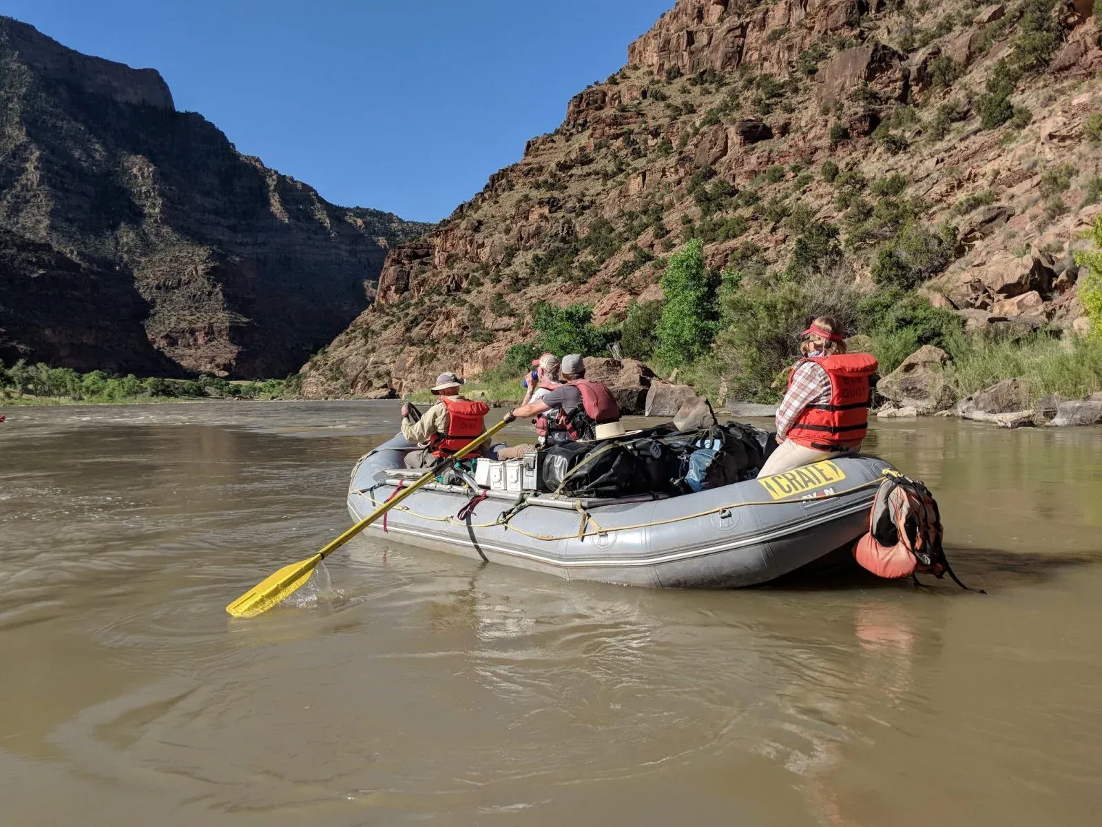 Rafting on a muddy river through a canyon. People in orange vests are rowing.