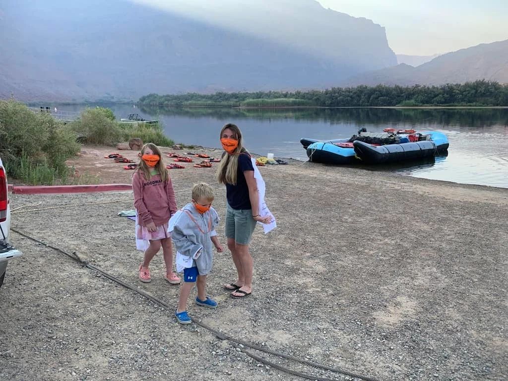 Three people wearing orange face coverings stand on a gravel beach by a river with a raft. Mountains in the background.
