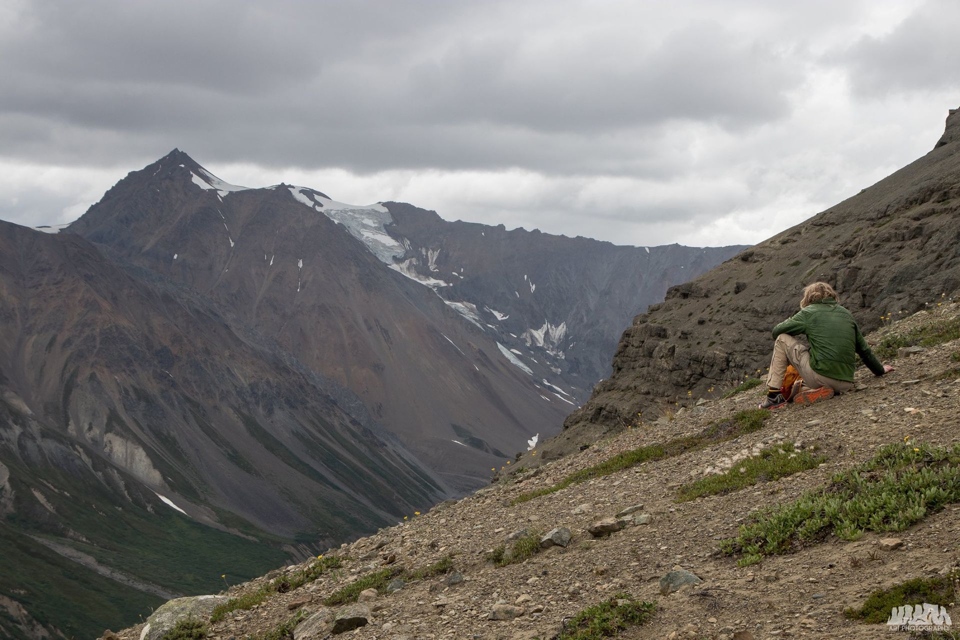 Person rests on rocky hillside, overlooking mountain valley under cloudy sky.