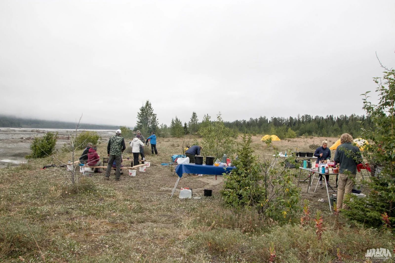 Group of people setting up camp on a grassy riverbank under an overcast sky.