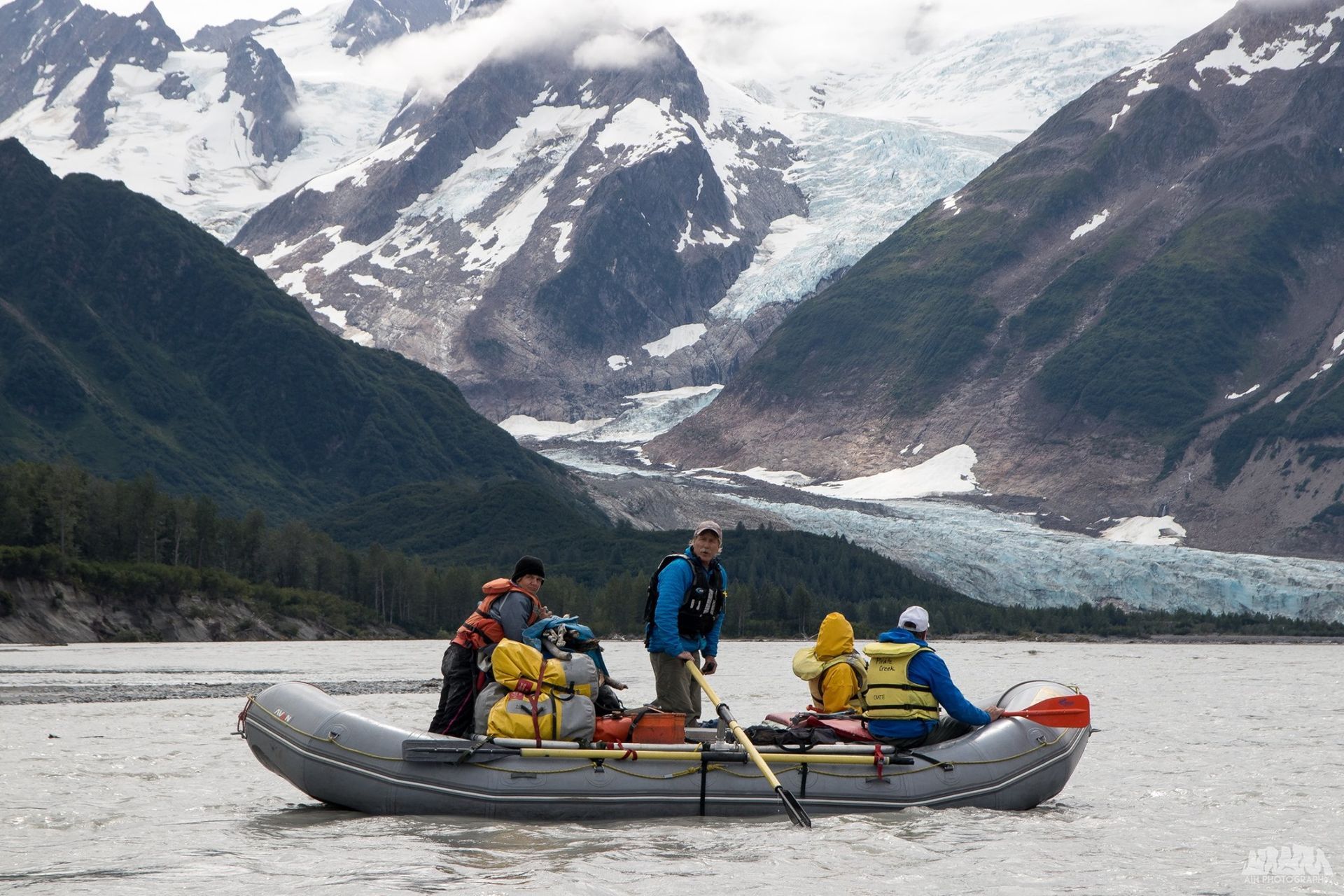 People on raft in glacial river, mountains and glacier in background.