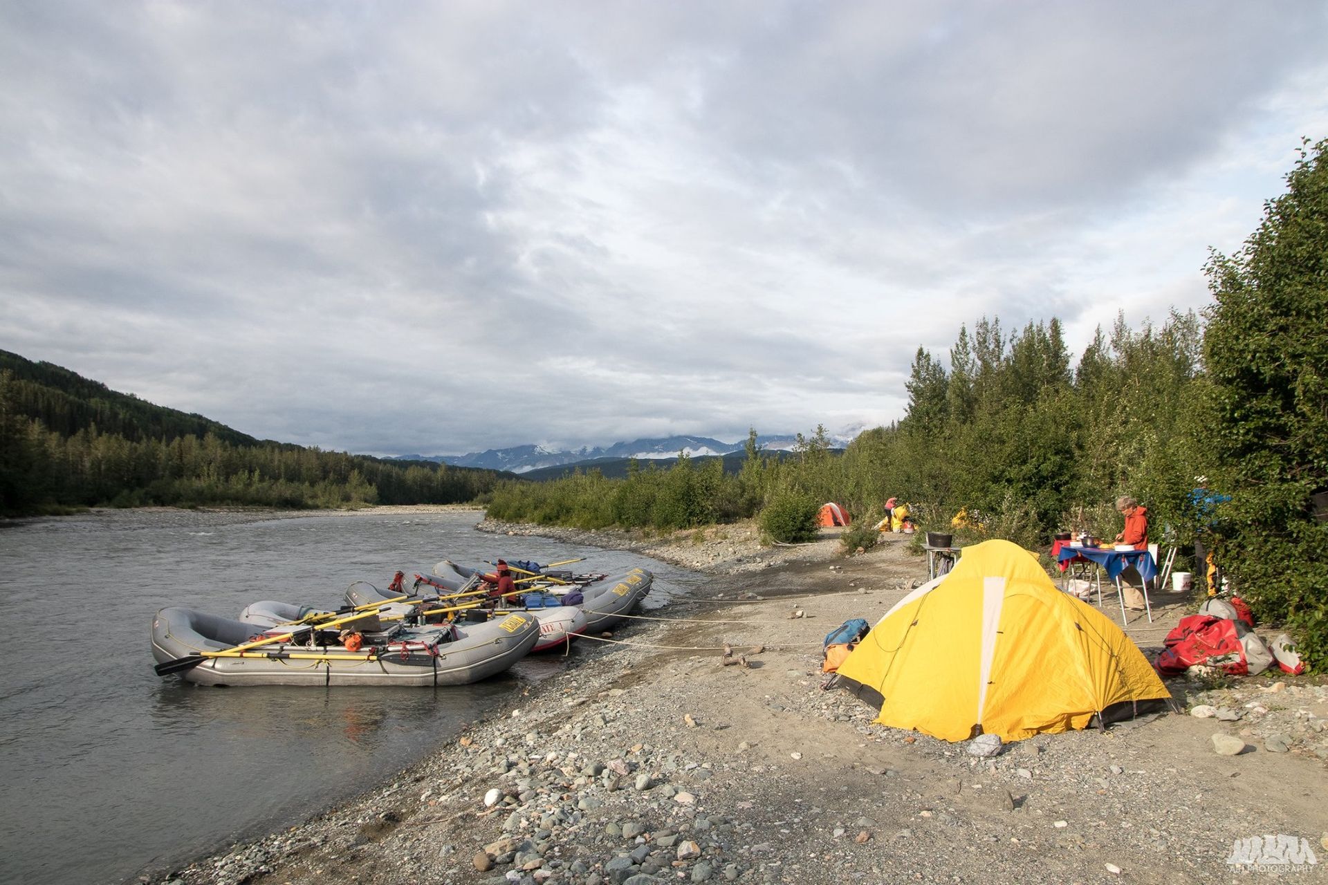 Yellow tent on gravel shore next to river, rafts, trees, and cloudy sky.